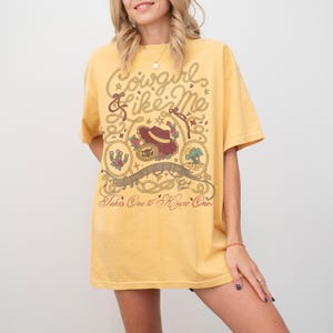 May include: A mustard yellow short-sleeve t-shirt featuring a brown graphic print with the text Cowgirl Like Me, Eyes full of Stars, and Takes One to Know One. The design includes a cowboy hat, a horseshoe, a cactus, and rope motifs.