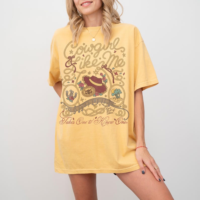 May include: A mustard yellow short-sleeve t-shirt featuring a brown graphic print with the text Cowgirl Like Me, Eyes full of Stars, and Takes One to Know One. The design includes a cowboy hat, a horseshoe, a cactus, and rope motifs.
