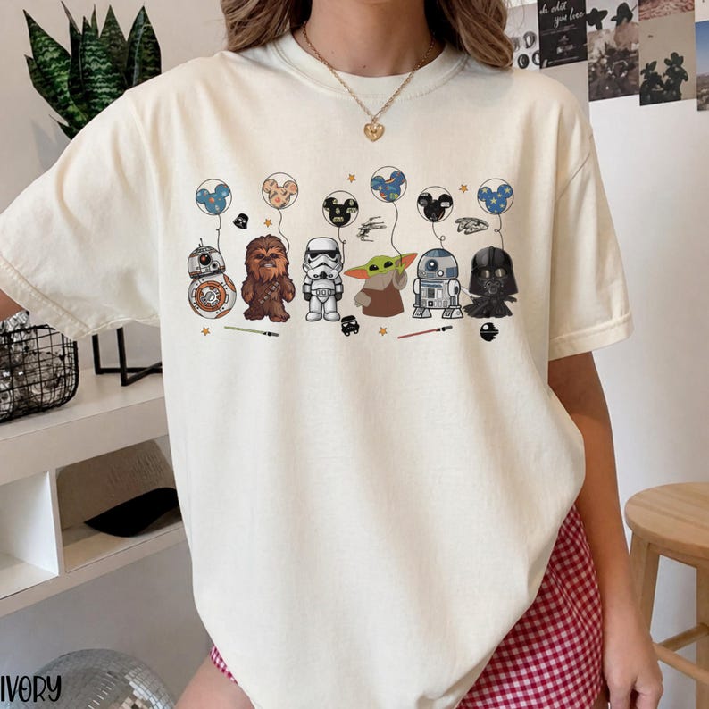 May include: Ivory-colored t-shirt featuring cartoon Star Wars characters holding Mickey Mouse-shaped balloons. Characters include BB-8, Chewbacca, a Stormtrooper, Grogu, R2-D2, and Darth Vader. The design includes stars and lightsabers.