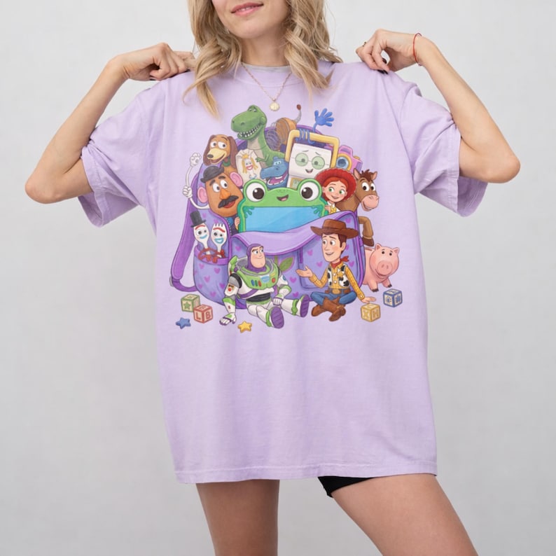 May include: A light purple t-shirt featuring a colorful graphic of various animated characters. The design includes a frog, a green dinosaur, a cowboy, a space ranger, and a pig. The shirt is a comfortable, oversized fit.