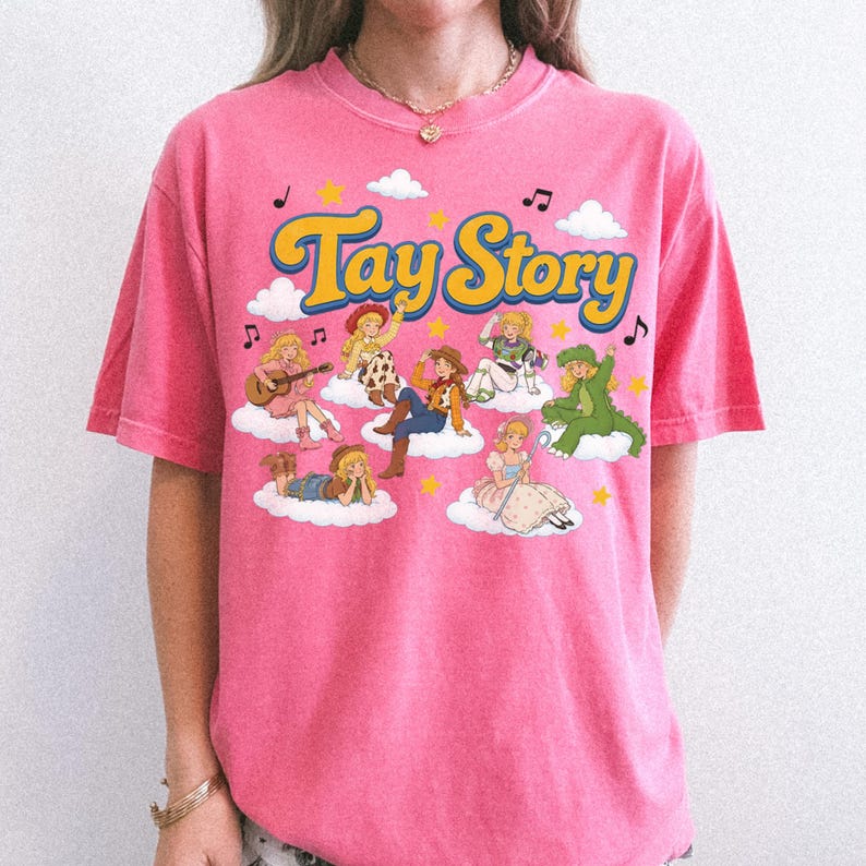 Puede incluir: Una camiseta rosa brillante de manga corta con el texto Tay Story en amarillo con bordes azules. El gr&aacute;fico incluye varias figuras ilustradas con diversos disfraces, situadas sobre nubes blancas con estrellas amarillas y notas musicales negras.