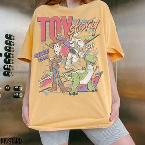 Pode incluir: Camiseta amarelo mostarda com o logotipo Toy Story e os personagens Woody, Buzz Lightyear e Rex. O gr&aacute;fico inclui o texto "Toy Story" em rosa e "Since 1995".