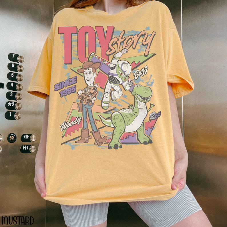 Pode incluir: Camiseta amarelo mostarda com o logotipo Toy Story e os personagens Woody, Buzz Lightyear e Rex. O gr&aacute;fico inclui o texto "Toy Story" em rosa e "Since 1995".