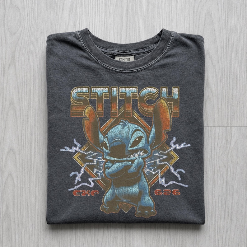 May include: Dark gray t-shirt featuring a graphic of the character Stitch from Lilo & Stitch. The design includes the word "STITCH" in a retro font, Stitch with crossed arms, lightning bolts, and the number "626".