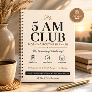 Könnte beinhalten: Ein cremefarbener Spiralplaner mit dem Text "5 AM CLUB" und "Morning Routine Planner". Das Cover hat eine goldfarbene Spiralbindung. Der Planer enthält Abschnitte für einen Tagesplaner, einen Gewohnheitstracker und ein 90-Tage-Protokoll. Ein goldener Stift liegt auf der Oberfläche.