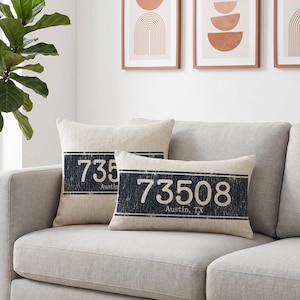 Pode incluir: Duas almofadas bege com um design azul marinho desgastado que se assemelha a uma placa de carro. Uma almofada diz "735 Austin" e a outra "73508 Austin, TX". As almofadas estão em um sofá cinza.
