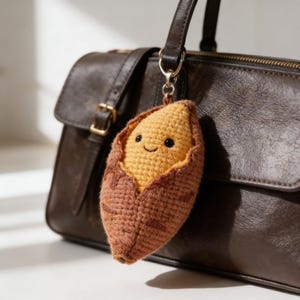 May include: A crocheted sweet potato keychain with a smiling face, in shades of brown and orange. It hangs from a silver-coloured keyring. The background shows a brown leather handbag.