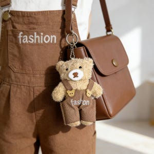 May include: A brown teddy bear keyring wearing brown corduroy dungarees, with the word "fashion" embroidered on the front. The bear hangs from a brown leather bag. The word "fashion" is also embroidered on the brown dungarees.