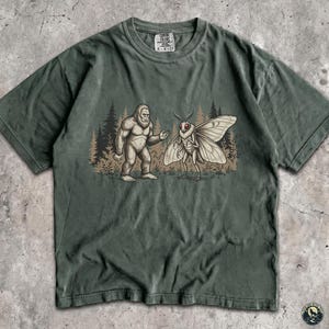 May include: A moss green short-sleeve t-shirt featuring a graphic illustration of a Bigfoot standing next to a large moth in a forest setting. The shirt has a crew neckline and a visible brand label on the inside of the collar area.