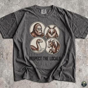 Può includere: Una t-shirt a maniche corte grigio antracite con una stampa grafica di quattro illustrazioni circolari raffiguranti un Bigfoot, una creatura simile a una falena, un serpente e una capra alata. Sotto i cerchi, il testo recita RESPECT THE LOCALS.