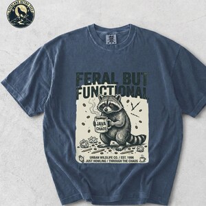 May include: Denim blue t-shirt featuring a raccoon graphic holding a coffee mug. The text on the shirt reads "FERAL BUT FUNCTIONAL" and "JAVA CHAOS". Additional text includes "URBAN WILDLIFE CO. / EST. 1996 JUST HOWLING / THROUGH THE CHAOS".