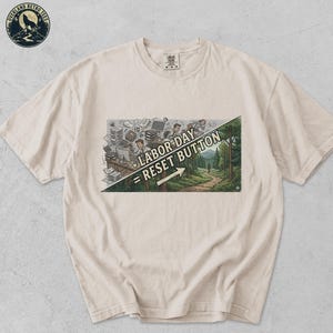 May include: An ivory short-sleeve t-shirt featuring a graphic print divided diagonally. The left side shows a cluttered office with a person at a desk, while the right side depicts a forest path. Text reads LABOR DAY = RESET BUTTON with an arrow.