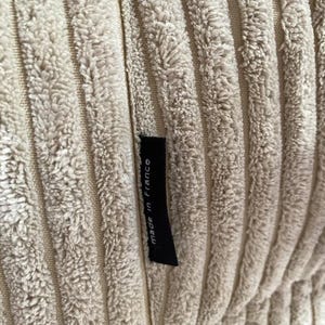 May include: Close-up of a beige corduroy fabric with vertical ridges. A small black tag is sewn into the fabric and reads "made in France". The fabric has a soft, textured appearance.