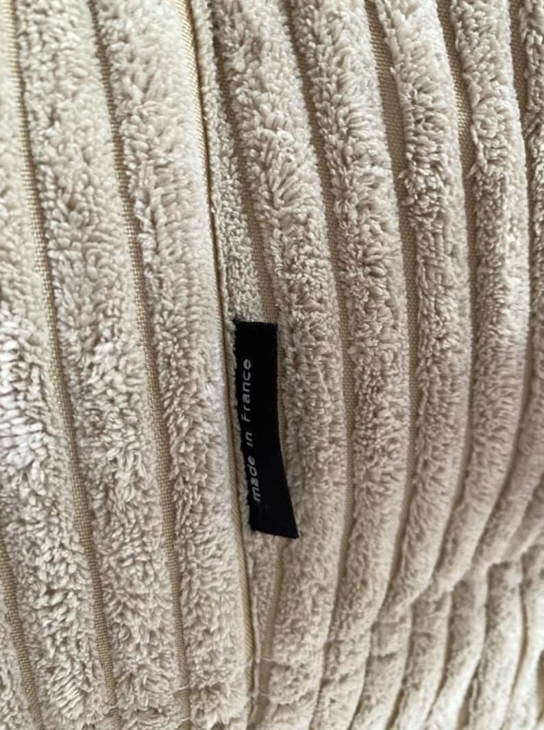 May include: Close-up of a beige corduroy fabric with vertical ridges. A small black tag is sewn into the fabric and reads "made in France". The fabric has a soft, textured appearance.