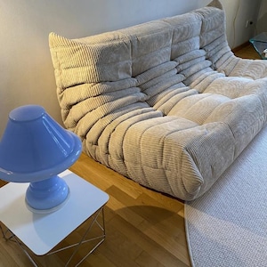 May include: A beige corduroy sofa with a low profile and rounded edges. A light blue lamp sits on a white side table with a metal frame. The room has hardwood floors and a light-colored rug.