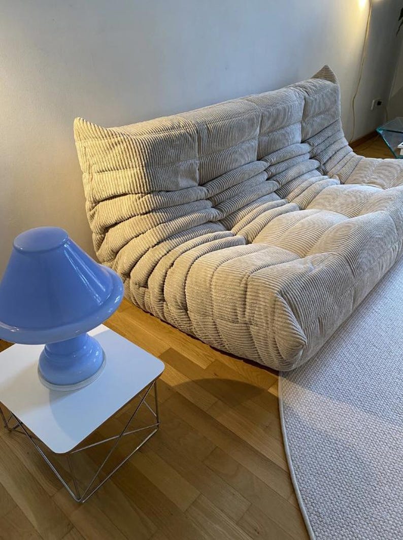 May include: A beige corduroy sofa with a low profile and rounded edges. A light blue lamp sits on a white side table with a metal frame. The room has hardwood floors and a light-colored rug.