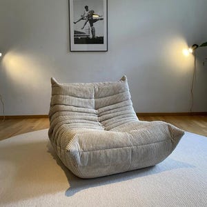 May include: A beige corduroy lounge chair with a low profile and rounded edges. The chair is placed on a light-colored rug. A framed black and white photograph hangs on the wall behind the chair.