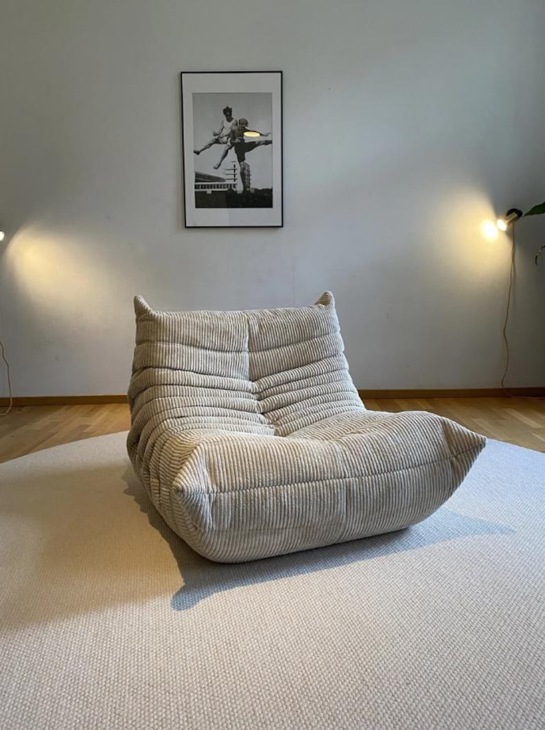May include: A beige corduroy lounge chair with a low profile and rounded edges. The chair is placed on a light-colored rug. A framed black and white photograph hangs on the wall behind the chair.
