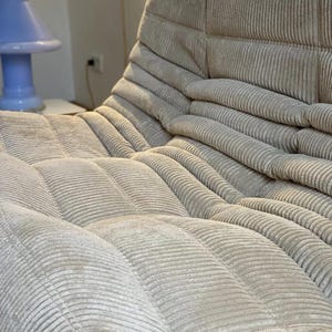 May include: A beige corduroy sofa with a low profile and a unique, modular design. The sofa features a series of cushioned sections, creating a comfortable and inviting seating area. The corduroy fabric has a soft texture and a subtle sheen.