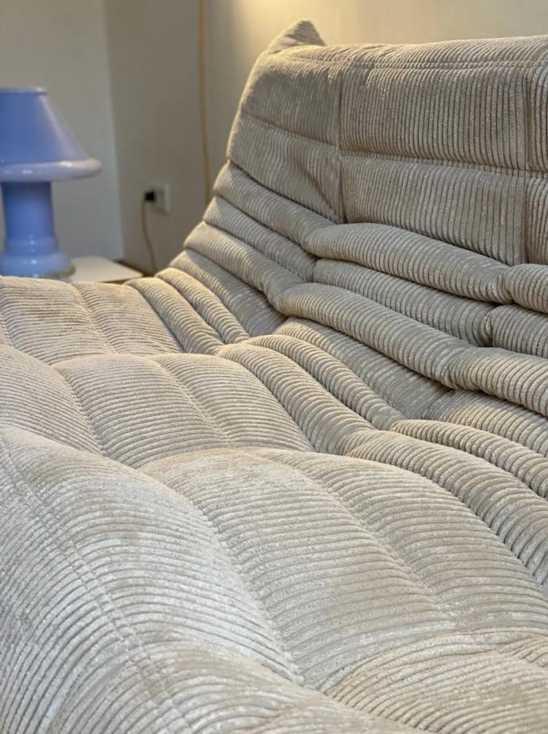 May include: A beige corduroy sofa with a low profile and a unique, modular design. The sofa features a series of cushioned sections, creating a comfortable and inviting seating area. The corduroy fabric has a soft texture and a subtle sheen.