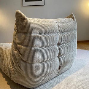 May include: A light beige corduroy lounge chair with a rounded back and a textured, ribbed design. The chair is on a light gray rug, and a framed black and white photograph hangs on the wall in the background.