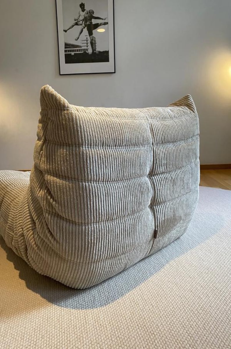 May include: A light beige corduroy lounge chair with a rounded back and a textured, ribbed design. The chair is on a light gray rug, and a framed black and white photograph hangs on the wall in the background.