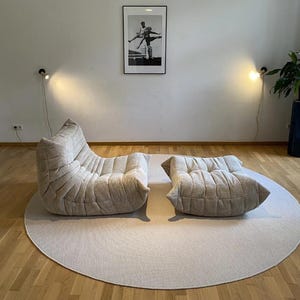 May include: A beige corduroy lounge chair and ottoman set on a round, light-colored rug. The chair has a unique, curved design. A framed black and white photograph hangs on the wall. Two wall-mounted lamps provide soft lighting.