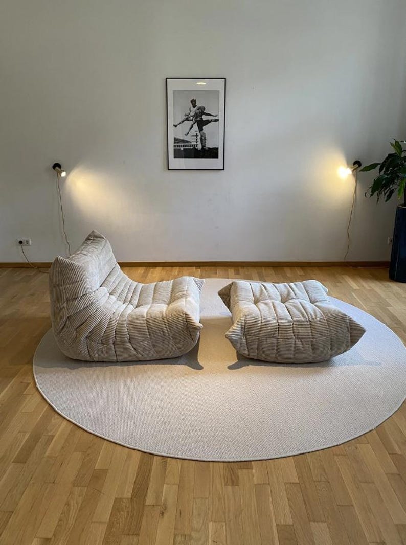 May include: A beige corduroy lounge chair and ottoman set on a round, light-colored rug. The chair has a unique, curved design. A framed black and white photograph hangs on the wall. Two wall-mounted lamps provide soft lighting.