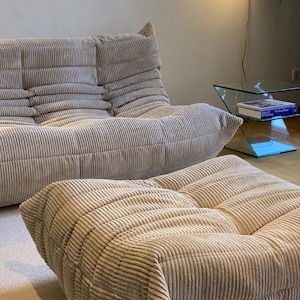 May include: Beige corduroy sofa and ottoman set. The sofa has a low profile with a multi-cushion design. The ottoman is square and matches the sofa's corduroy fabric. The furniture is in a well-lit room with a glass side table.