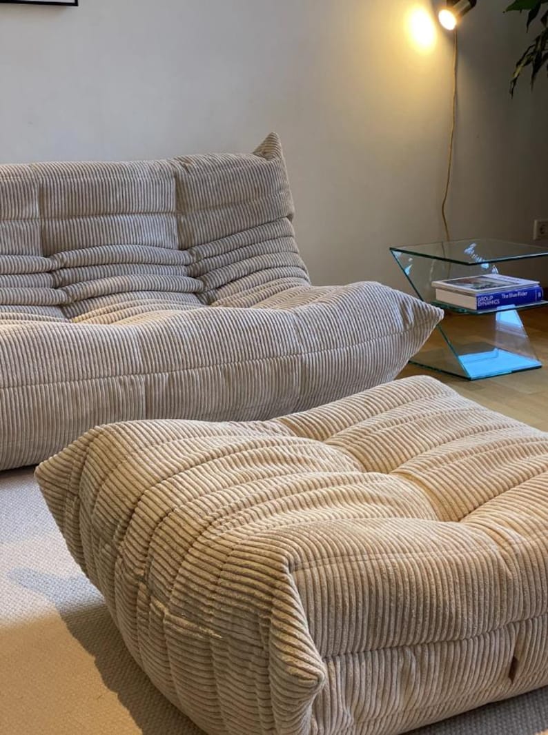 May include: Beige corduroy sofa and ottoman set. The sofa has a low profile with a multi-cushion design. The ottoman is square and matches the sofa's corduroy fabric. The furniture is in a well-lit room with a glass side table.