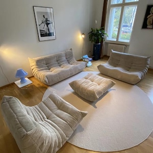 May include: A living room arrangement featuring beige corduroy upholstered modular sofas and a matching ottoman. The furniture sits on a light-colored round rug, with a hardwood floor. A blue lamp and artwork are also visible.