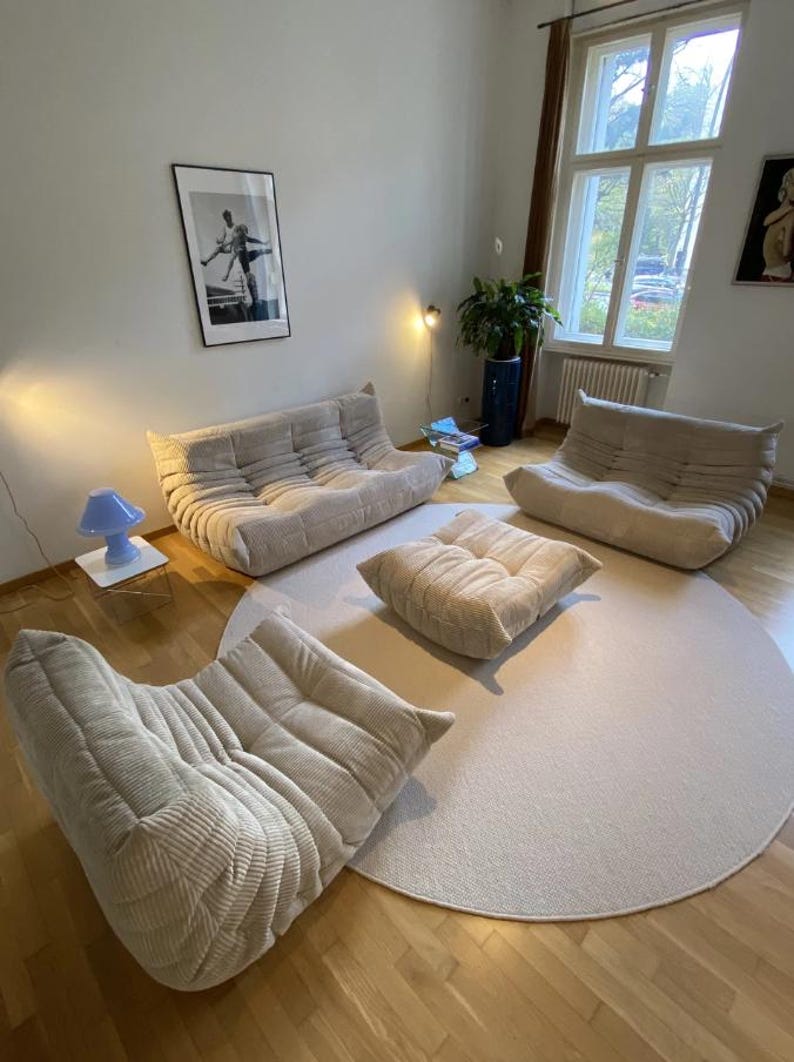 May include: A living room arrangement featuring beige corduroy upholstered modular sofas and a matching ottoman. The furniture sits on a light-colored round rug, with a hardwood floor. A blue lamp and artwork are also visible.