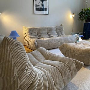 May include: Beige corduroy sofa and two matching ottomans in a living room setting. A blue lamp and a framed black and white photograph are also visible. The furniture is arranged on a light-colored rug.