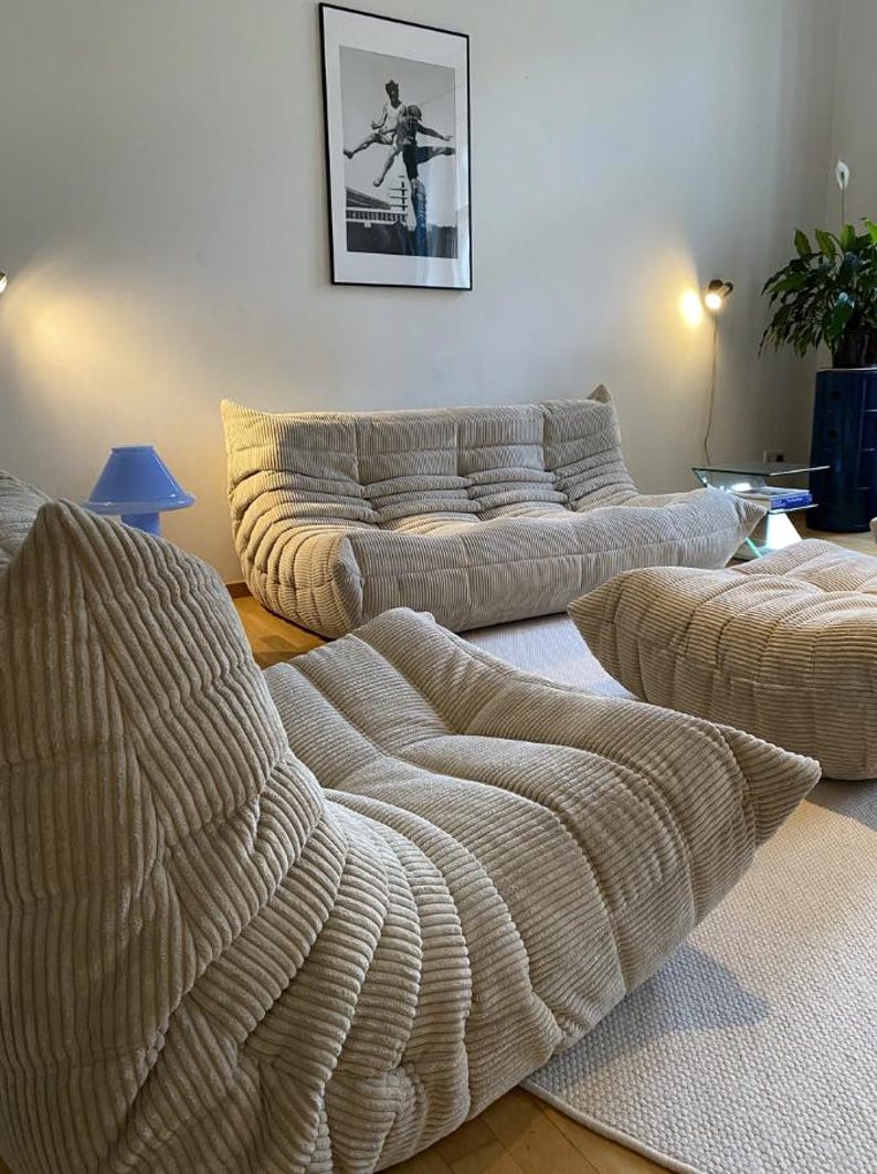 May include: Beige corduroy sofa and two matching ottomans in a living room setting. A blue lamp and a framed black and white photograph are also visible. The furniture is arranged on a light-colored rug.
