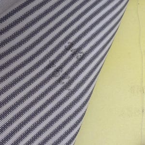 May include: Close-up of a fabric with a striped pattern in shades of gray and white. The fabric is angled, revealing a section of yellow material in the background. The stripes run diagonally across the fabric.