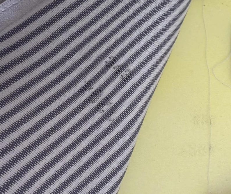 May include: Close-up of a fabric with a striped pattern in shades of gray and white. The fabric is angled, revealing a section of yellow material in the background. The stripes run diagonally across the fabric.