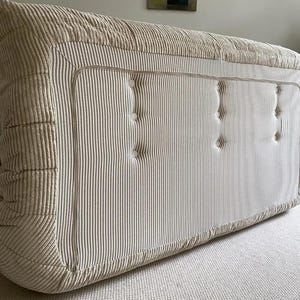 May include: A large, rectangular, beige corduroy ottoman with vertical striped detailing. The ottoman features a button-tufted design and a contrasting trim. The piece is placed on a neutral-colored carpet.