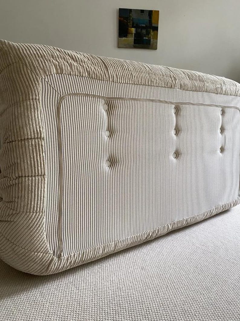 May include: A large, rectangular, beige corduroy ottoman with vertical striped detailing. The ottoman features a button-tufted design and a contrasting trim. The piece is placed on a neutral-colored carpet.