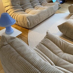 May include: Beige corduroy modular sofa with a matching ottoman and pillows. A light blue mushroom-shaped lamp sits on a white side table. A neutral-toned rug and hardwood flooring complete the living room setting.