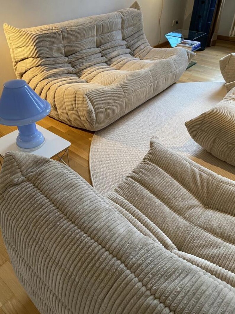 May include: Beige corduroy modular sofa with a matching ottoman and pillows. A light blue mushroom-shaped lamp sits on a white side table. A neutral-toned rug and hardwood flooring complete the living room setting.