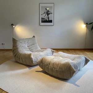May include: A beige corduroy armchair and ottoman set sit on a round, light-colored rug. The furniture is in a modern style. A black and white framed picture hangs on the wall behind the furniture. Two wall-mounted lamps are on either side of the picture.