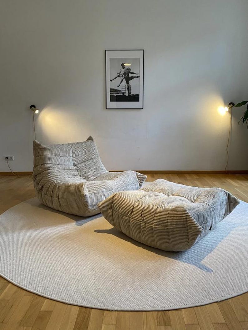 May include: A beige corduroy armchair and ottoman set sit on a round, light-colored rug. The furniture is in a modern style. A black and white framed picture hangs on the wall behind the furniture. Two wall-mounted lamps are on either side of the picture.