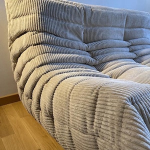 May include: A beige corduroy sofa with a distinctive, rounded design. The sofa features a series of cushioned sections, creating a textured, inviting appearance. The corduroy fabric has a ribbed pattern, adding depth and visual interest to the furniture.