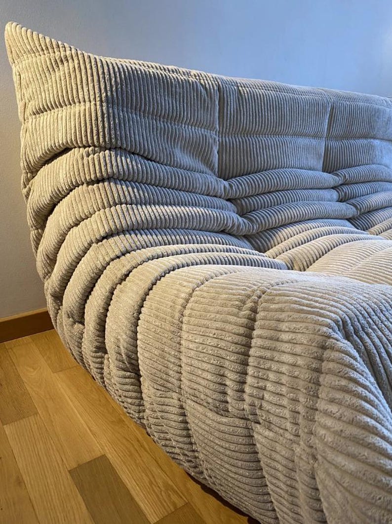 May include: A beige corduroy sofa with a distinctive, rounded design. The sofa features a series of cushioned sections, creating a textured, inviting appearance. The corduroy fabric has a ribbed pattern, adding depth and visual interest to the furniture.