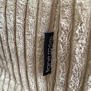May include: Close-up of a beige corduroy fabric with vertical ridges. A black tag with the white text "ligne roset" is sewn into the fabric. The fabric has a soft, textured appearance.