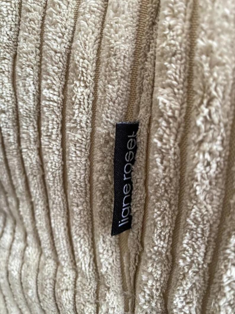 May include: Close-up of a beige corduroy fabric with vertical ridges. A black tag with the white text "ligne roset" is sewn into the fabric. The fabric has a soft, textured appearance.