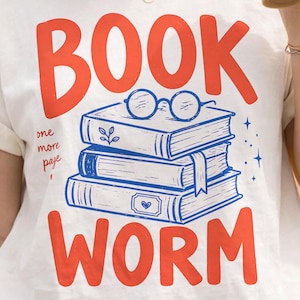 以下が含まれることがあります： 赤い文字でBOOK WORMと書かれ、丸い眼鏡が乗った3冊の本の積み重ねが青いグラフィックで描かれた白いTシャツ。本の横には小さな青い筆記体でone more pageと書かれており、本には葉やハートのアイコンがあしらわれています。