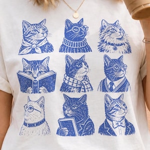 May include: A white short-sleeve t-shirt featuring a blue graphic print of nine distinct cats arranged in a grid. Each cat is depicted with different accessories, including scarves, glasses, sweaters, and books, rendered in a detailed illustrative style.