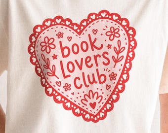 May include: A cream-colored short-sleeve t-shirt featuring a large red heart graphic on the chest. The heart has a scalloped lace-like border and contains the text book lovers club in a red script font, surrounded by small floral and heart motifs.