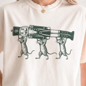 May include: A cream-coloured short-sleeve t-shirt featuring a dark green graphic print of three mice standing on their hind legs, collectively lifting a large, detailed nutcracker figure horizontally above their heads.
